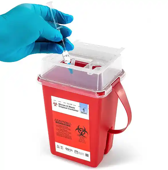 A gloved hand holding a syringe above a red biohazard waste disposal container with a transparent lid.